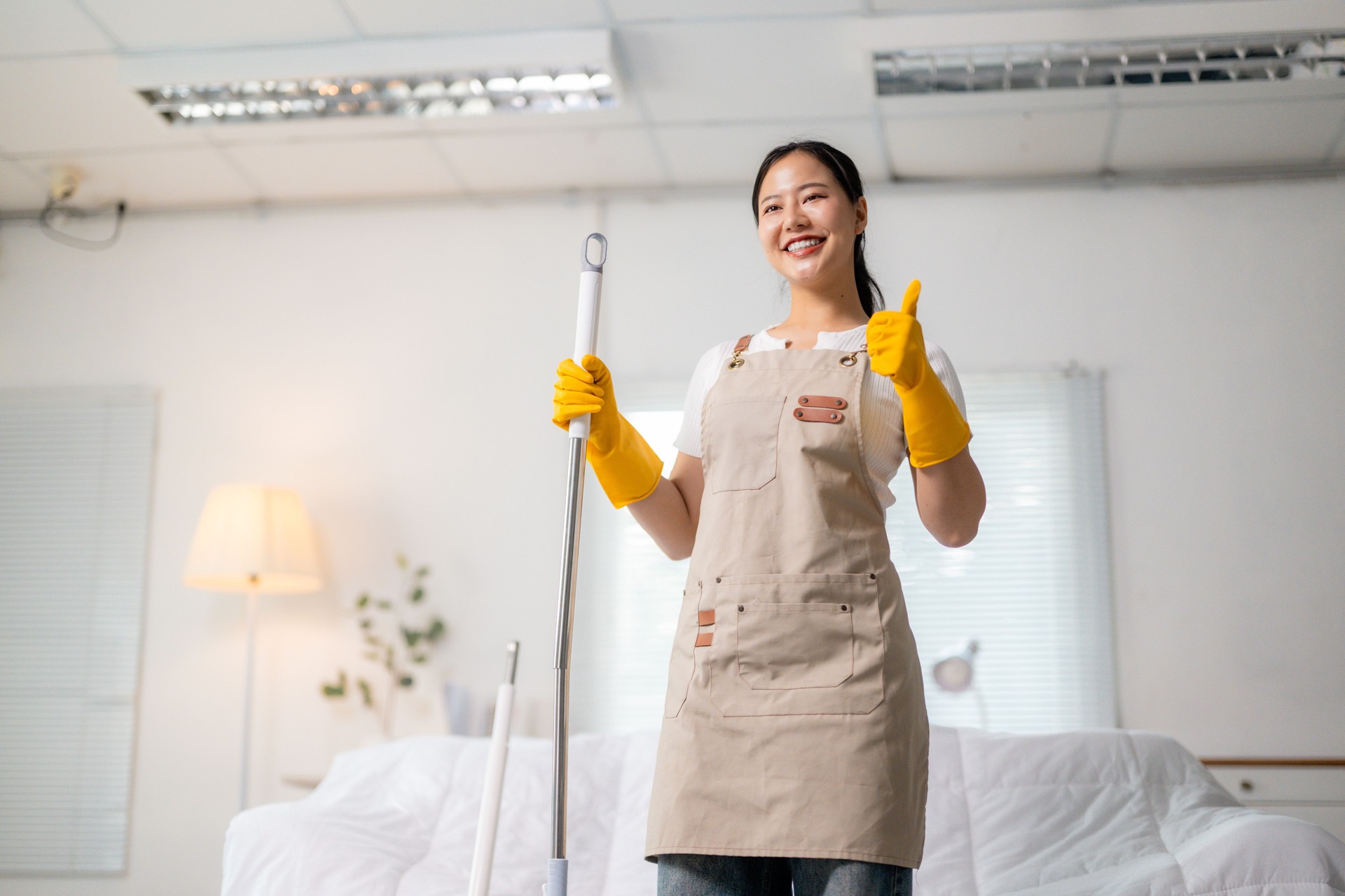 Asian woman wearing an apron and yellow gloves holds a mop and shows thumbs up, smiling at the camera